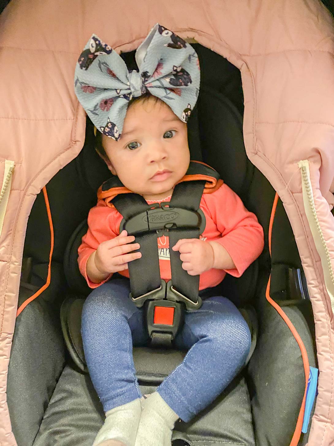 A sweet little Mosie baby girl with pink shirt and giant bow on her head sits in car seat