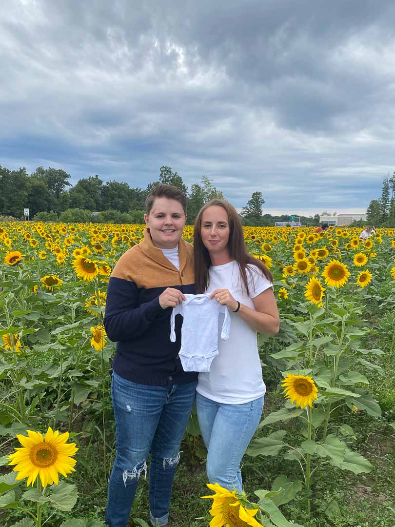 Two wives hold onesie together in sunflower field to announce their pregnancy!