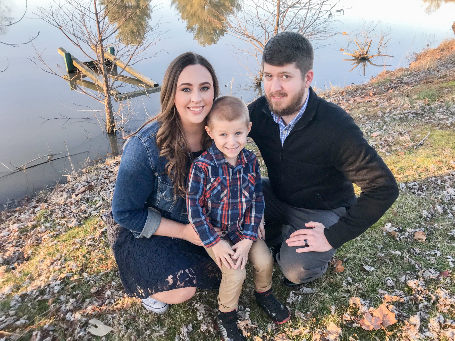 Family photo outside on fall day with Mom on the left kneeling in a denim jacket and long brown hair next to dad on the right with son sitting on their lap and all are smiling.