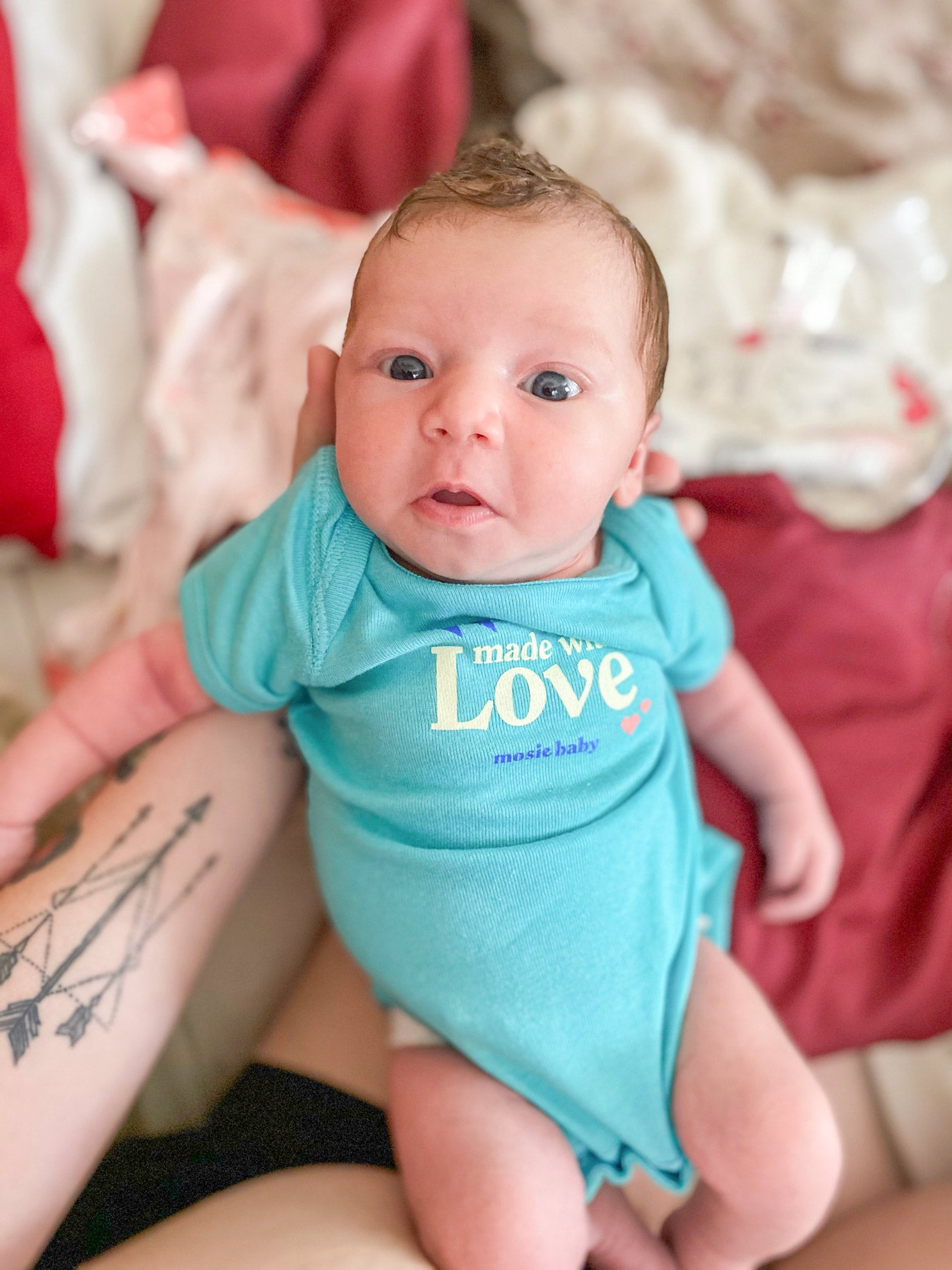 Newborn Mosie Baby in teal onsie being held up by momma's arms over a bed.