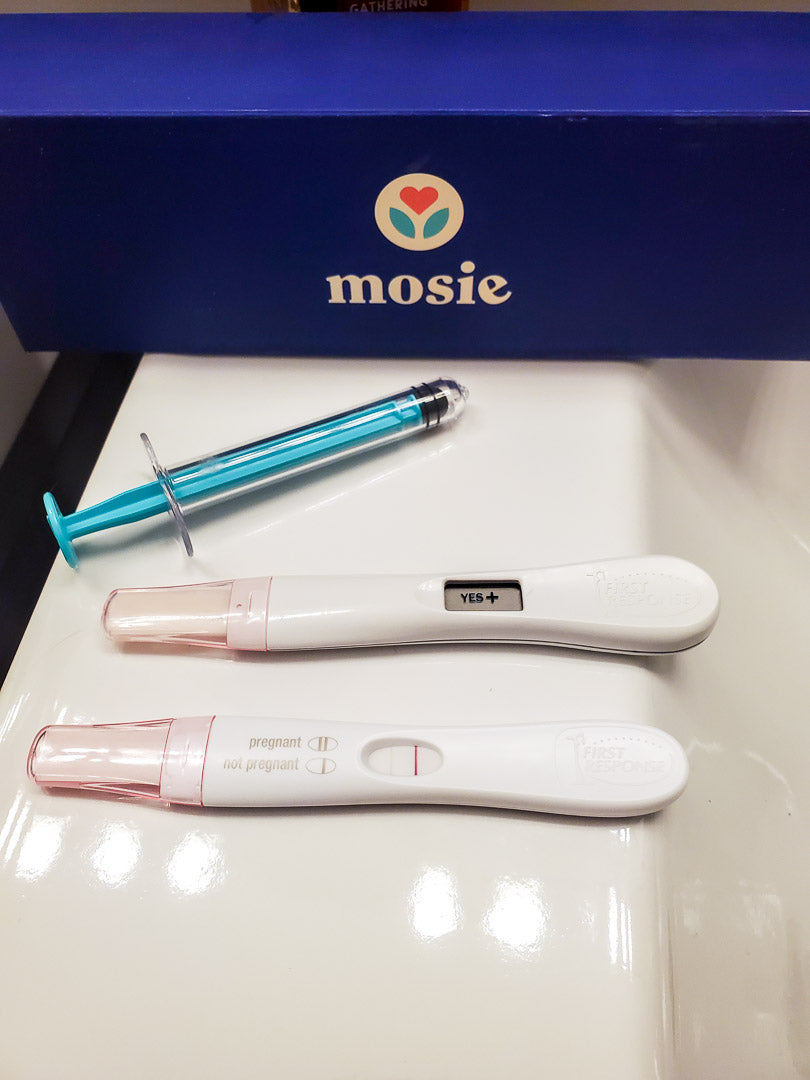 Mosie syringe and box laying next to positive pregnancy tests.