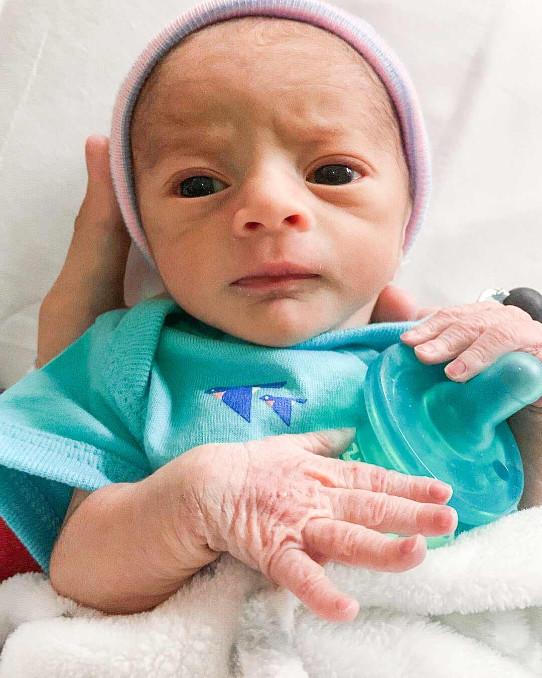 Mosie Baby newborn lays in NICU with onesie and hospital beanie celebrating release day from the hospital.